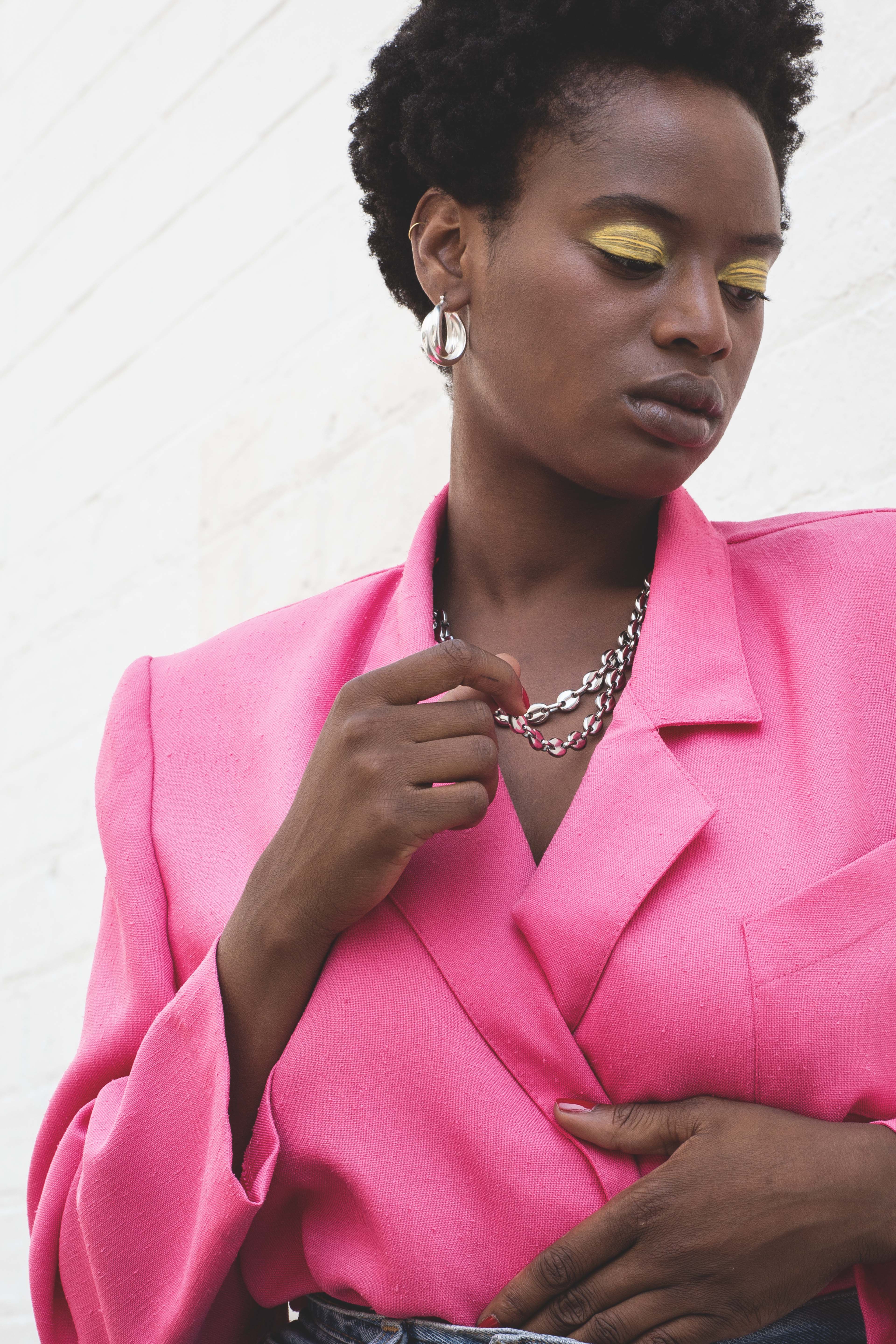 Femme en blazer rose vif, maquillage yeux jaunes, bijoux argentés, posant contre un mur blanc
