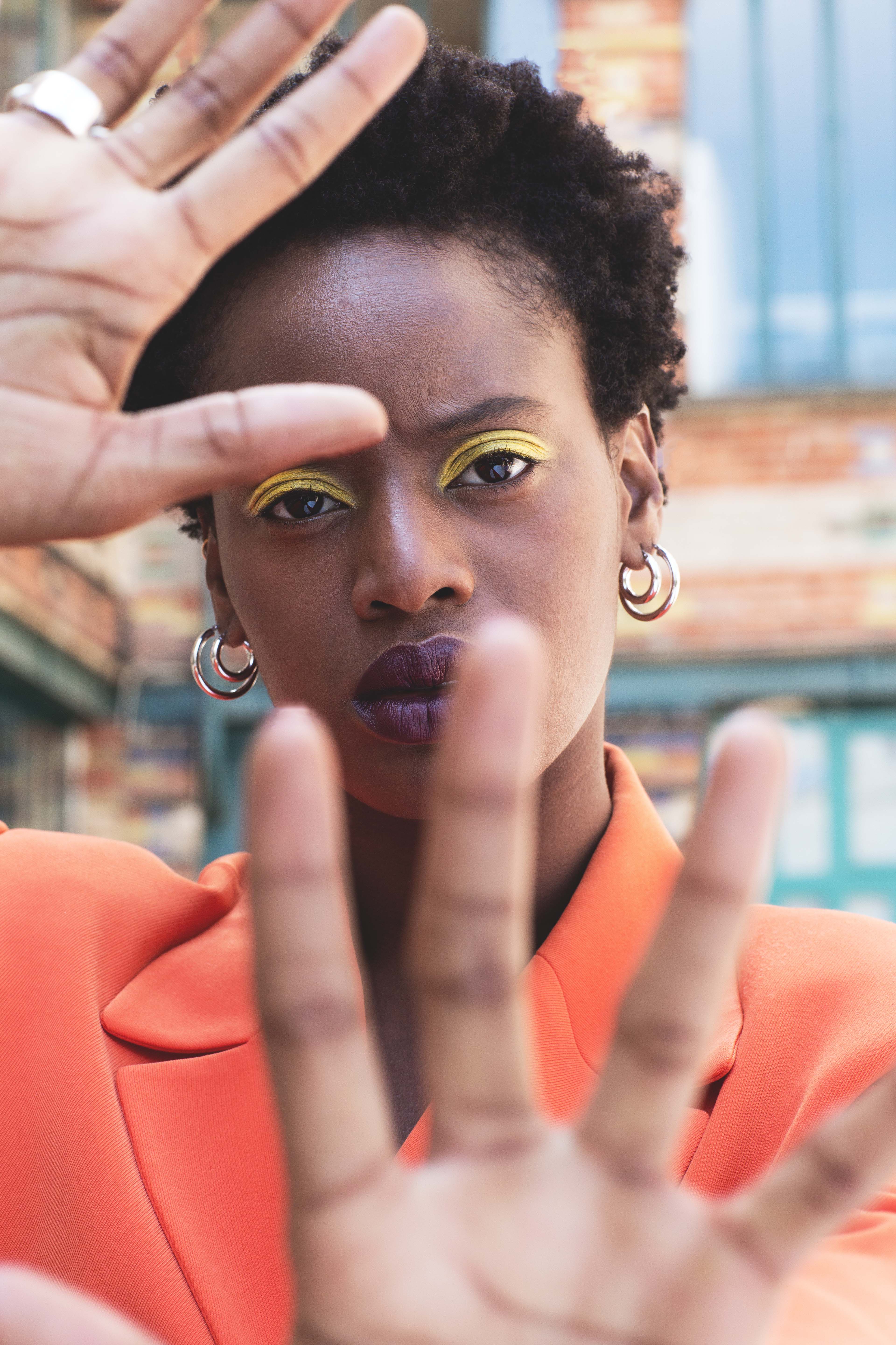 Portrait mode d’une femme portant un blazer orange, maquillage artistique yeux jaunes, main tendue vers l’objectif