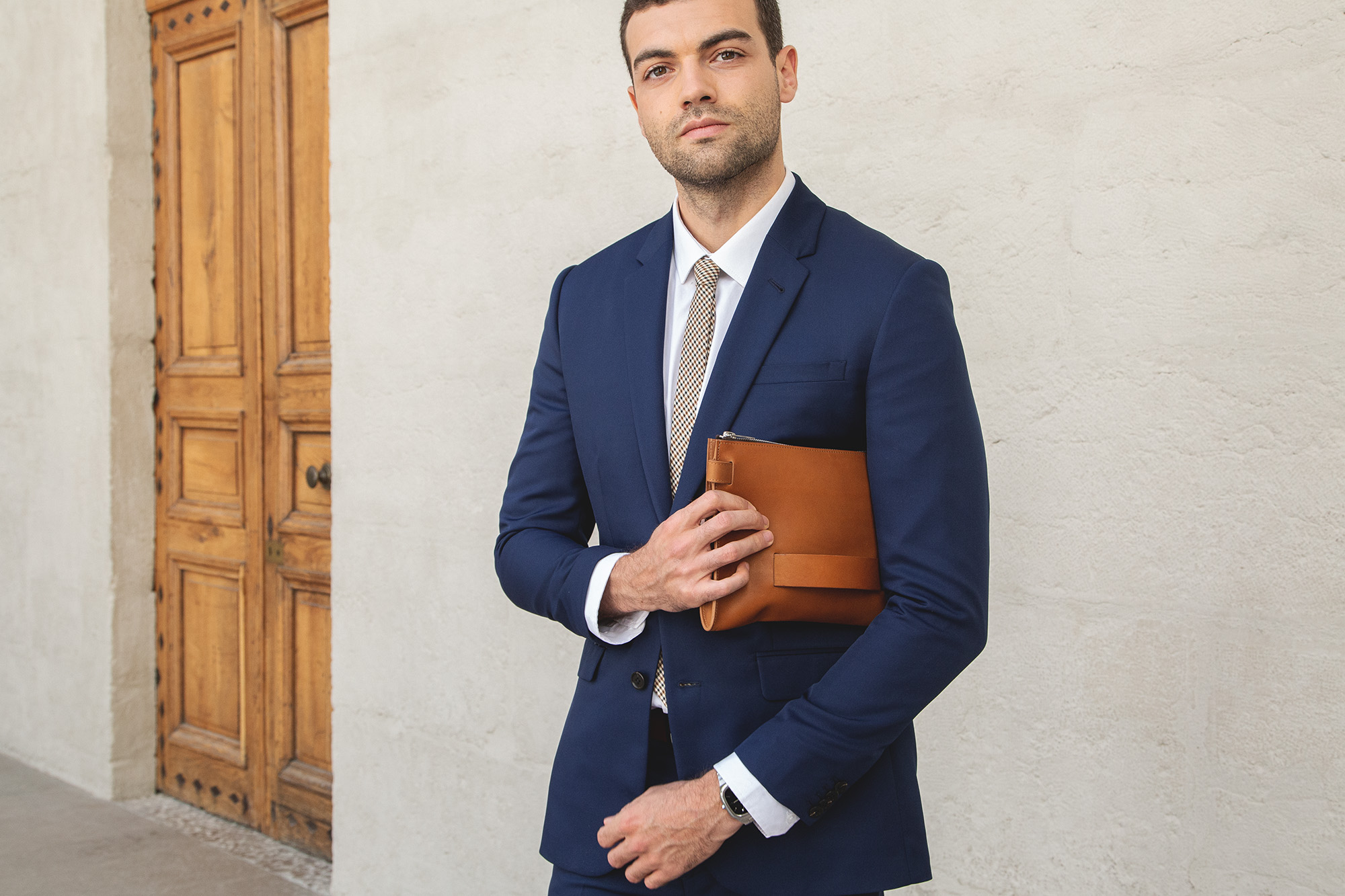 Homme en costume bleu marine avec chemise blanche, tenant une pochette marron, posé devant une porte en bois
