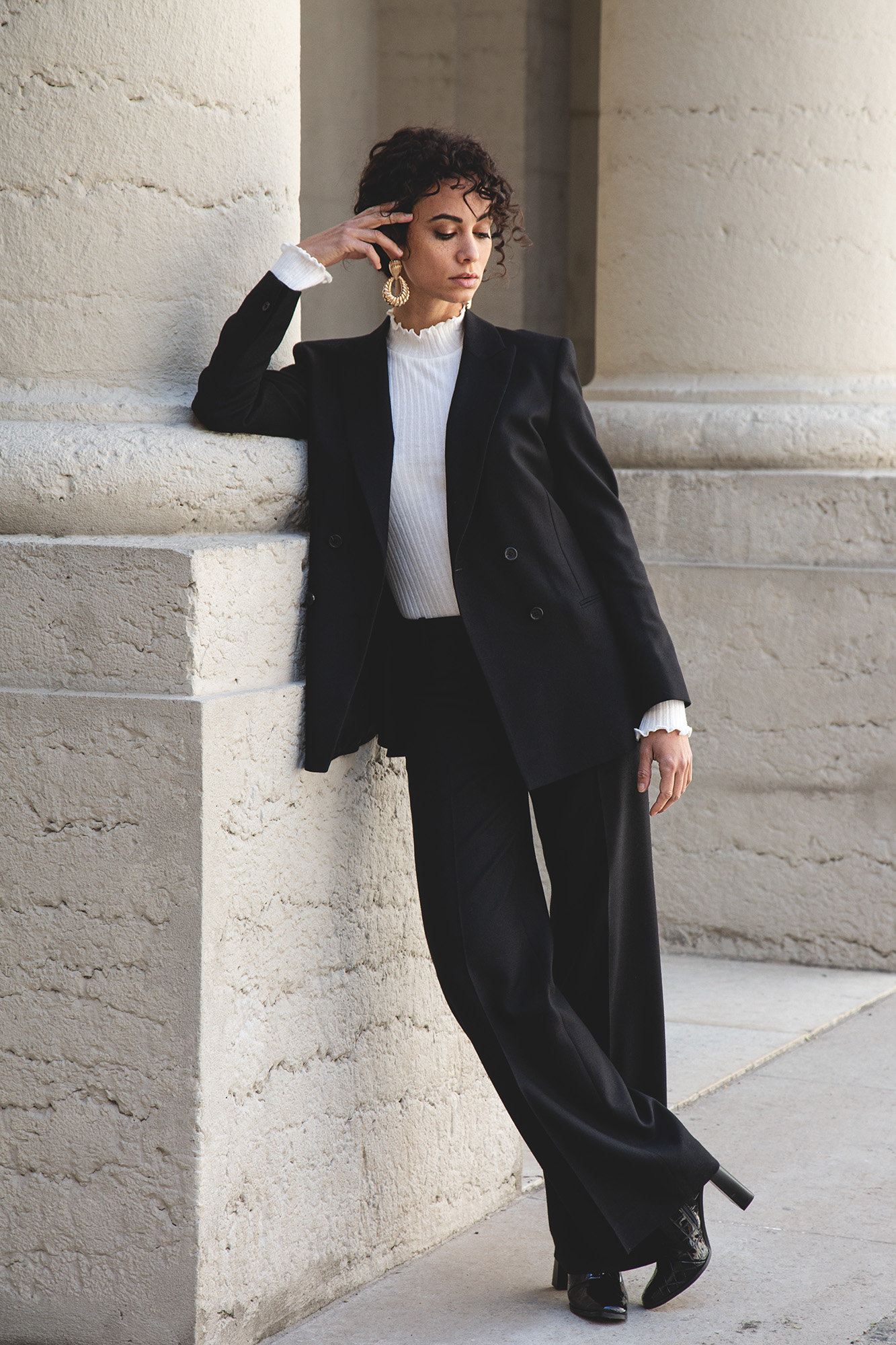 Femme en tailleur noir avec col roulé blanc, posant appuyée contre un mur en pierre