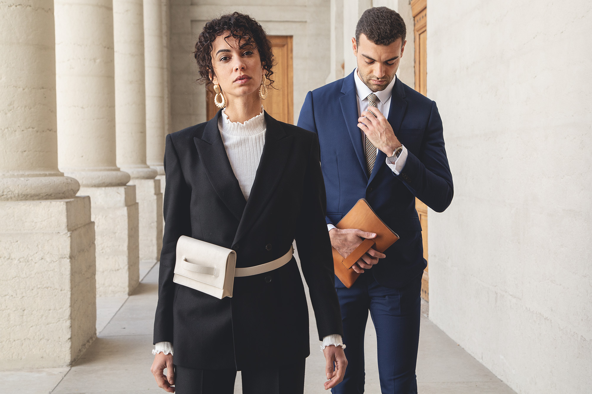 Une femme en tailleur noir et sac blanc aux côtés d'un homme en costume bleu tenant une pochette en cuir